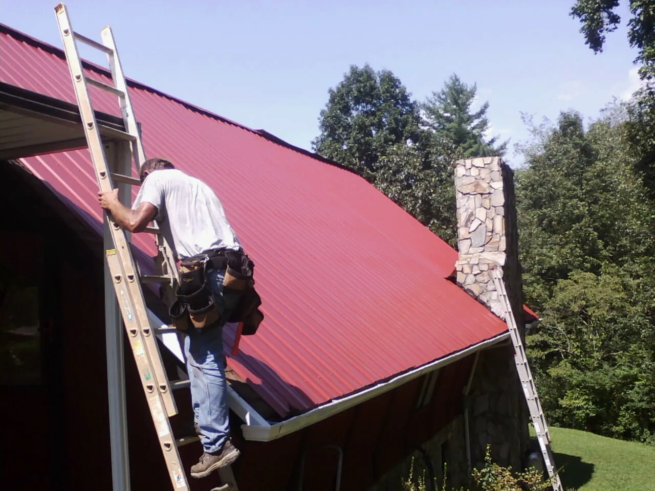 Roofer on a ladder beside a red metal roof and stone chimney — Soffit & Fascia Repair by Roofing Brothers in Northwood